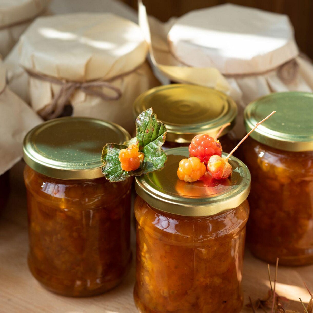 Jars of homemade cloudberry jam with golden-orange cloudberries on top, set on a wooden surface with rustic paper-wrapped jars in the background.
