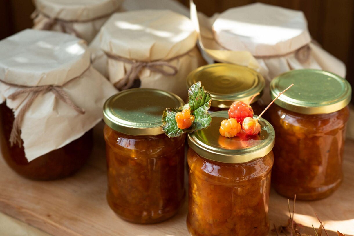 Jars of homemade cloudberry jam with golden-orange cloudberries on top, set on a wooden surface with rustic paper-wrapped jars in the background.