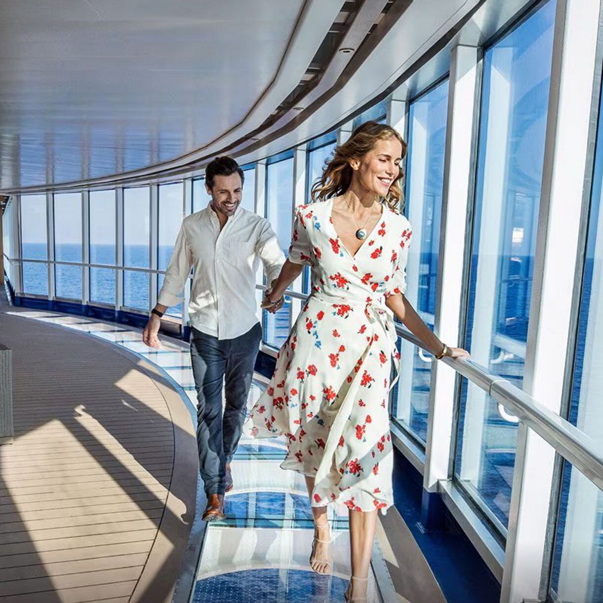 A couple holds hands and walks by the windows of their cruise ship.