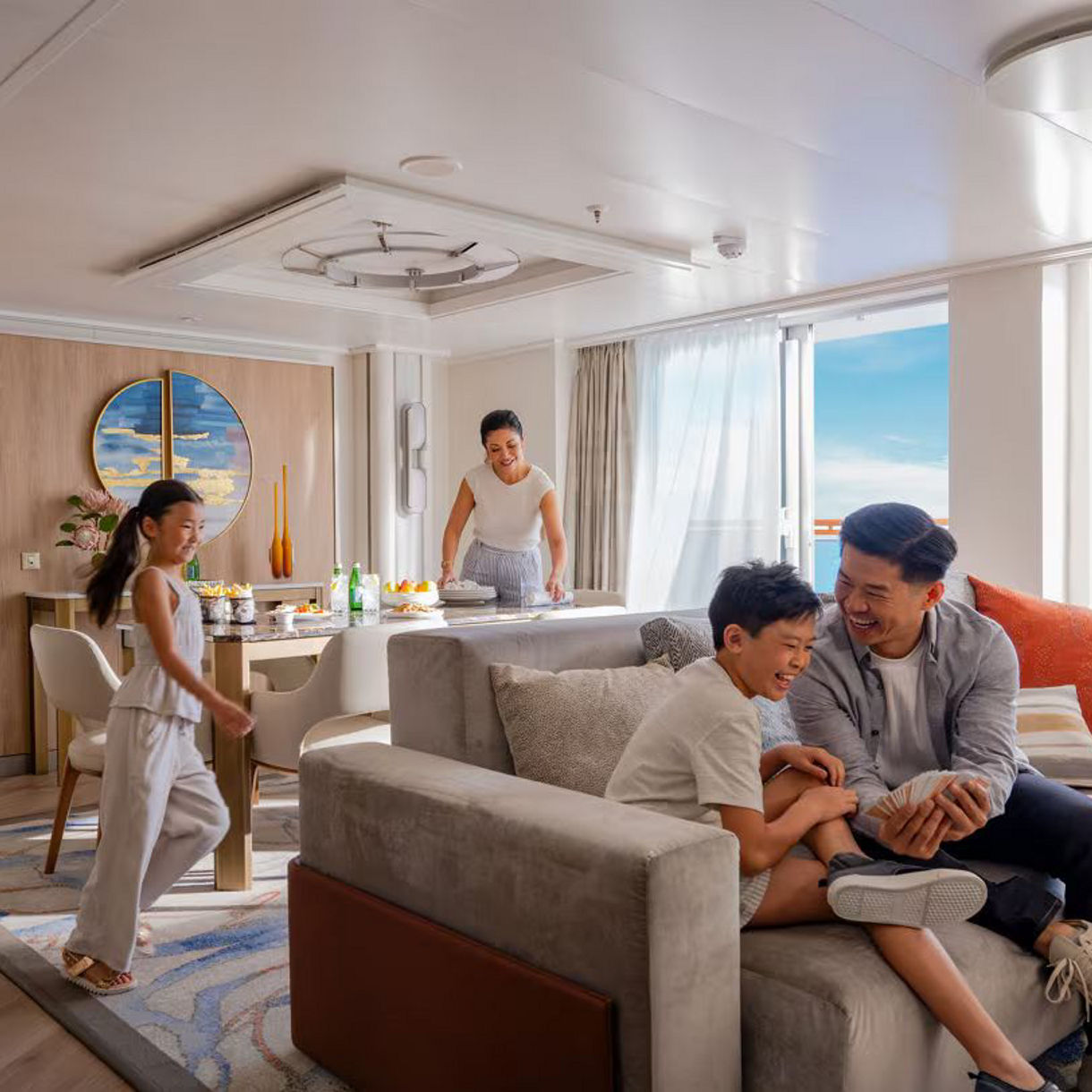 Family of four relaxing in a bright, modern cruise ship suite with large windows, comfortable seating area, and contemporary furnishings.