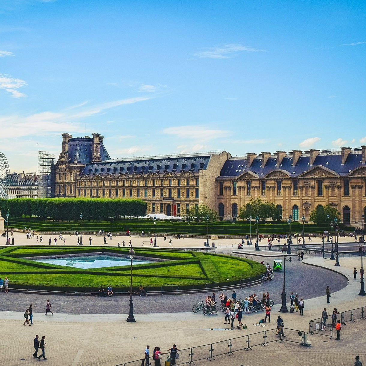 Aerial view of the Louvre Museum and Tuileries Garden in Paris, France, featuring the grand palace architecture, manicured circular garden and crowds of visitors under a blue sky with white clouds.