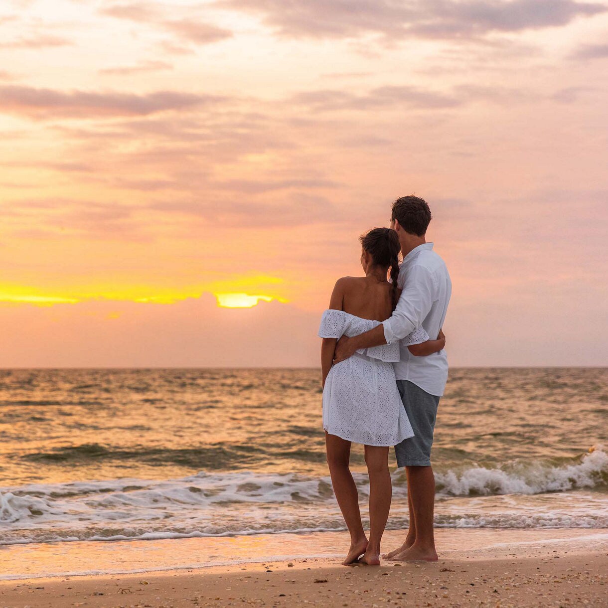 A couple dressed in white stands barefoot on a beach at sunset, gazing at the ocean as waves gently roll in.