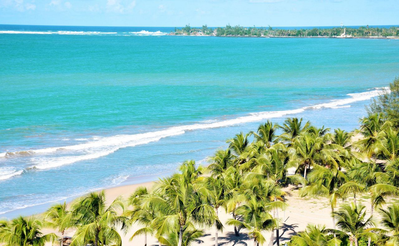 Aerial view of Isla Verde Beach in San Juan, Puerto Rico, showing palm trees, golden sand and turquoise ocean with buildings in the background.