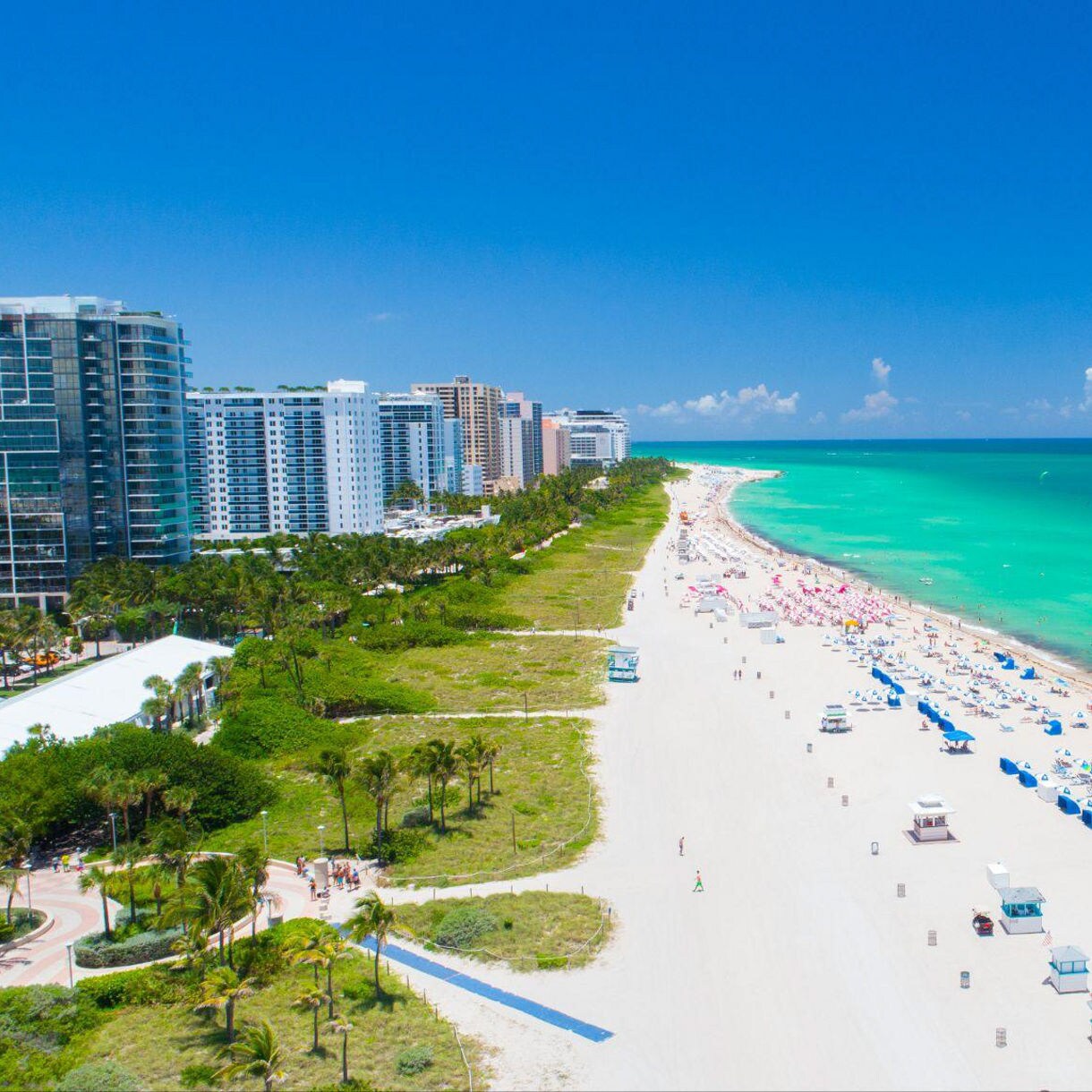 Guests stroll along South Beach in Miami, with lifeguard towers and umbrellas dotting the shore and turquoise waves rolling in under a bright blue sky.