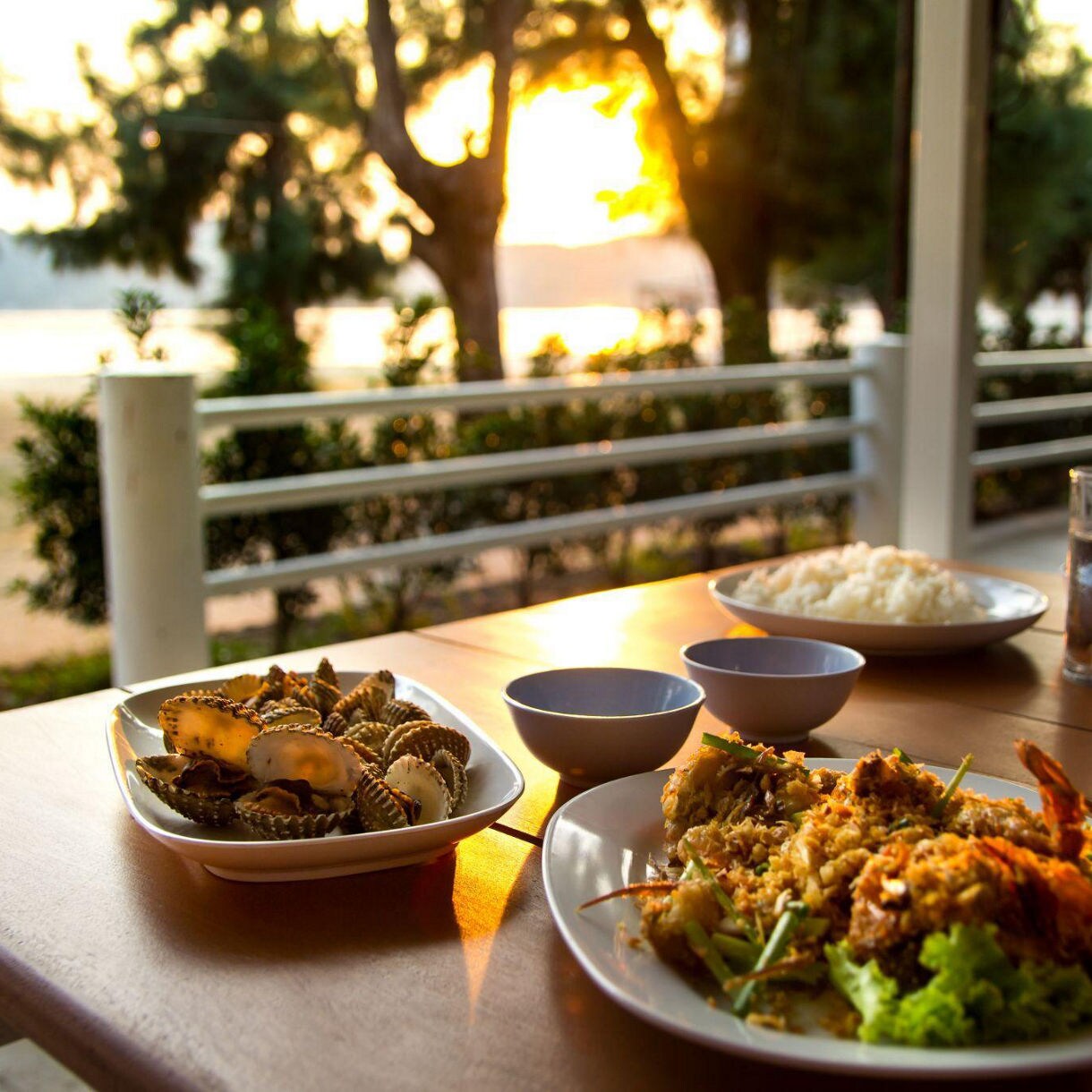 A table set with seafood dishes, rice, and drinks is placed outdoors overlooking a serene beach during sunset. The vibrant colors of the food contrast beautifully with the warm golden hues of the evening light. The setting evokes a relaxed and tranquil mood, perfect for enjoying a meal by the water.