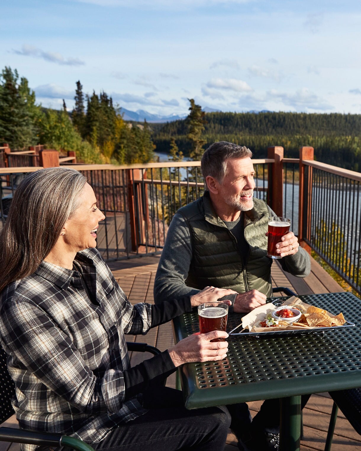 Two adults are seated at a green metal table on a deck overlooking a river and forested mountains. They are enjoying drinks and a meal in a relaxed, natural setting with clear skies and vibrant greenery. The scene conveys a peaceful, leisurely mood with casual attire and outdoor dining elements. The table features food and beverages, enhancing the sense of relaxation and enjoyment.