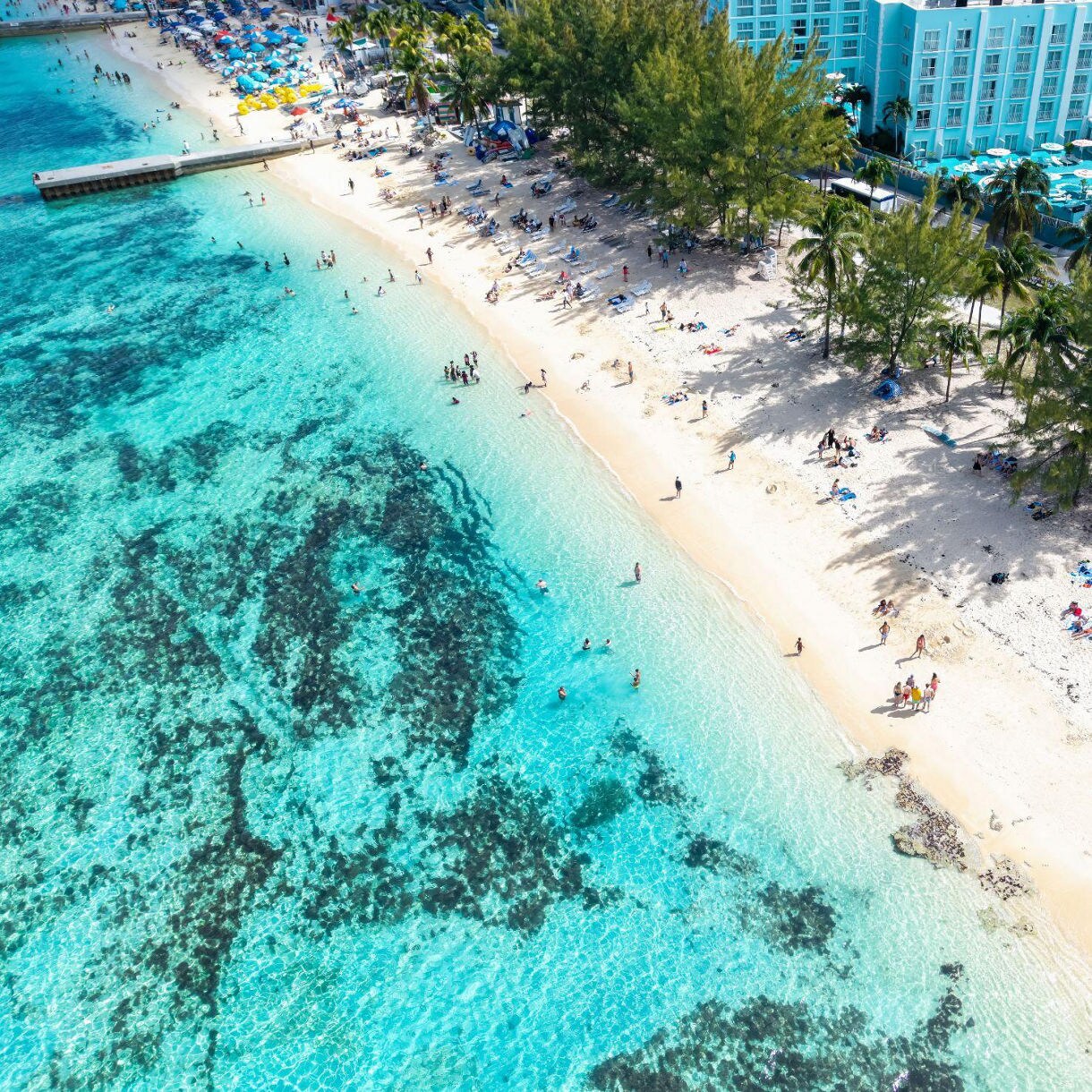 Tropical beach with two lounge chairs under a rainbow-colored umbrella, facing turquoise ocean water, with palm fronds and a small boat nearby.