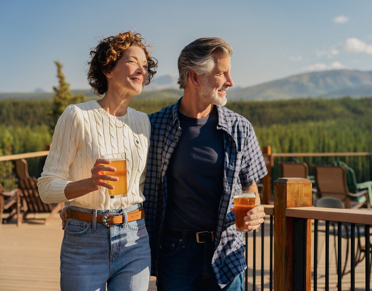 Two adults stand close together on a wooden deck, each holding a glass of beer. The setting overlooks a lush green forest and distant mountains under a clear blue sky. The atmosphere is relaxed and casual, with natural sunlight and outdoor furniture visible. Both individuals are dressed in casual attire, suggesting a leisurely outing.