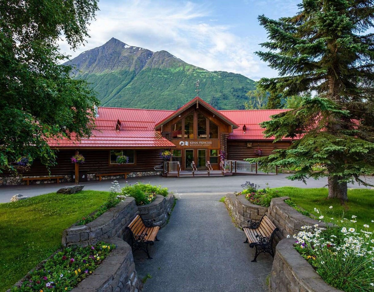 Front entrance of Kenai Princess Wilderness Lodge featuring log cabin construction with red metal roof, mountain peak backdrop, and landscaped courtyard with benches