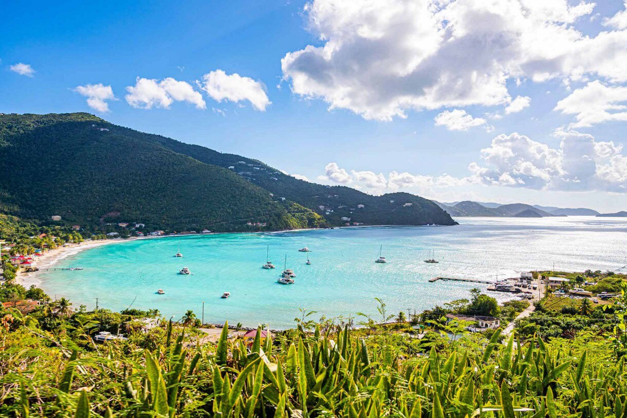 Scenic view of Cane Garden Bay in Tortola with turquoise water, anchored sailboats, green mountains, a beachside village, and partly cloudy skies.