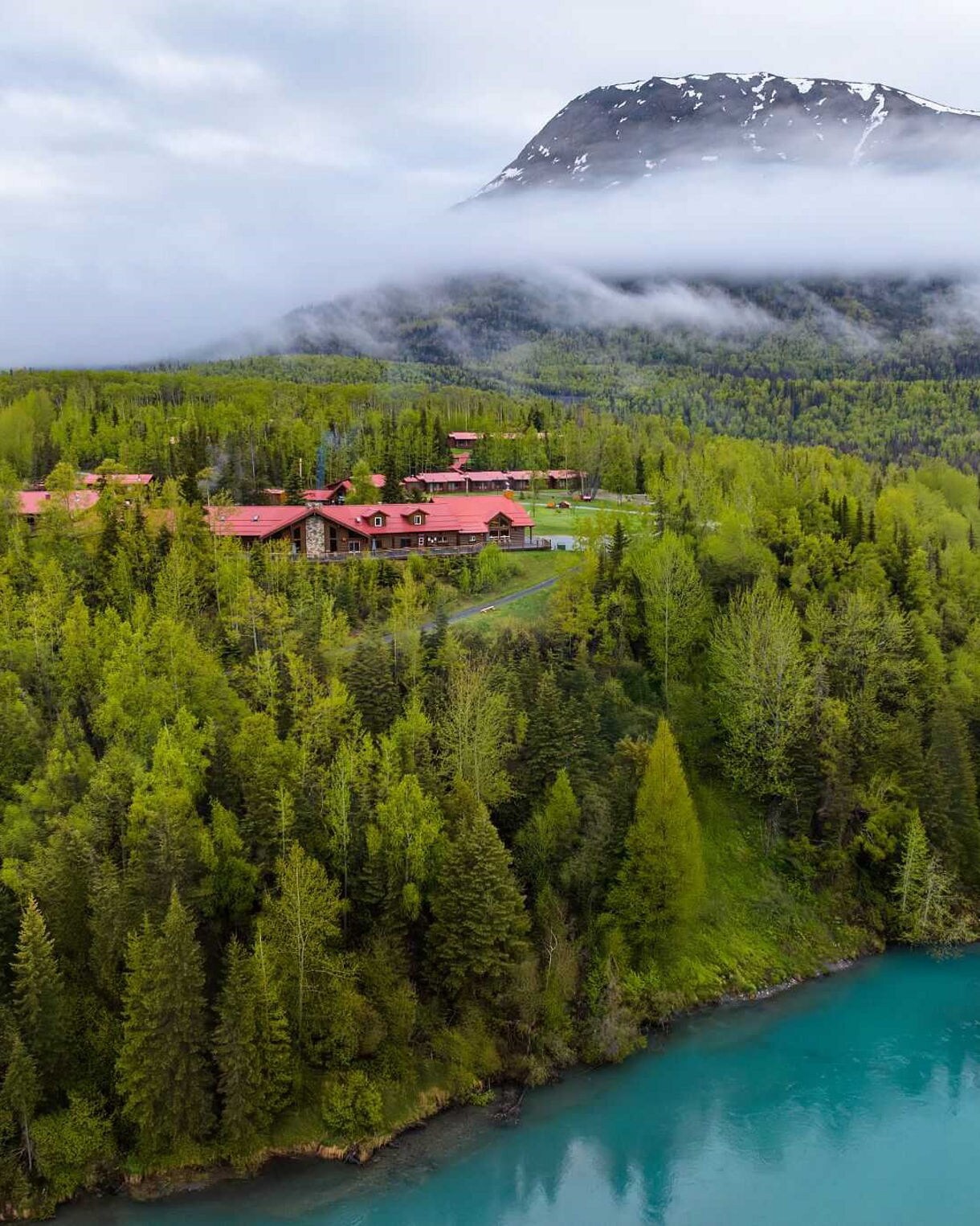 Aerial view of Kenai Princess Wilderness Lodge complex surrounded by dense evergreen forest with misty mountains in background and turquoise lake visible