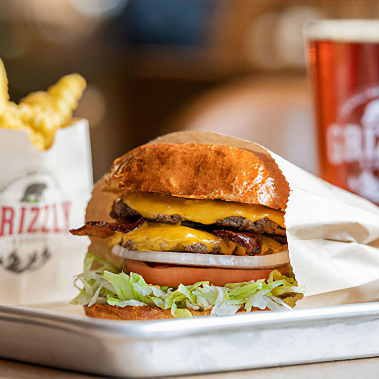 A cheeseburger with bacon, lettuce, tomato, and onion is served on a metal tray alongside crinkle-cut fries in a branded Grizzly bag. A beverage in a clear cup with the Grizzly logo is also visible in the background. The setting appears to be a casual dining restaurant with a warm, inviting atmosphere. The focus is on the fresh ingredients and appetizing presentation. The word 'GRIZZLY' is clearly visible on both the fry bag and drink cup.