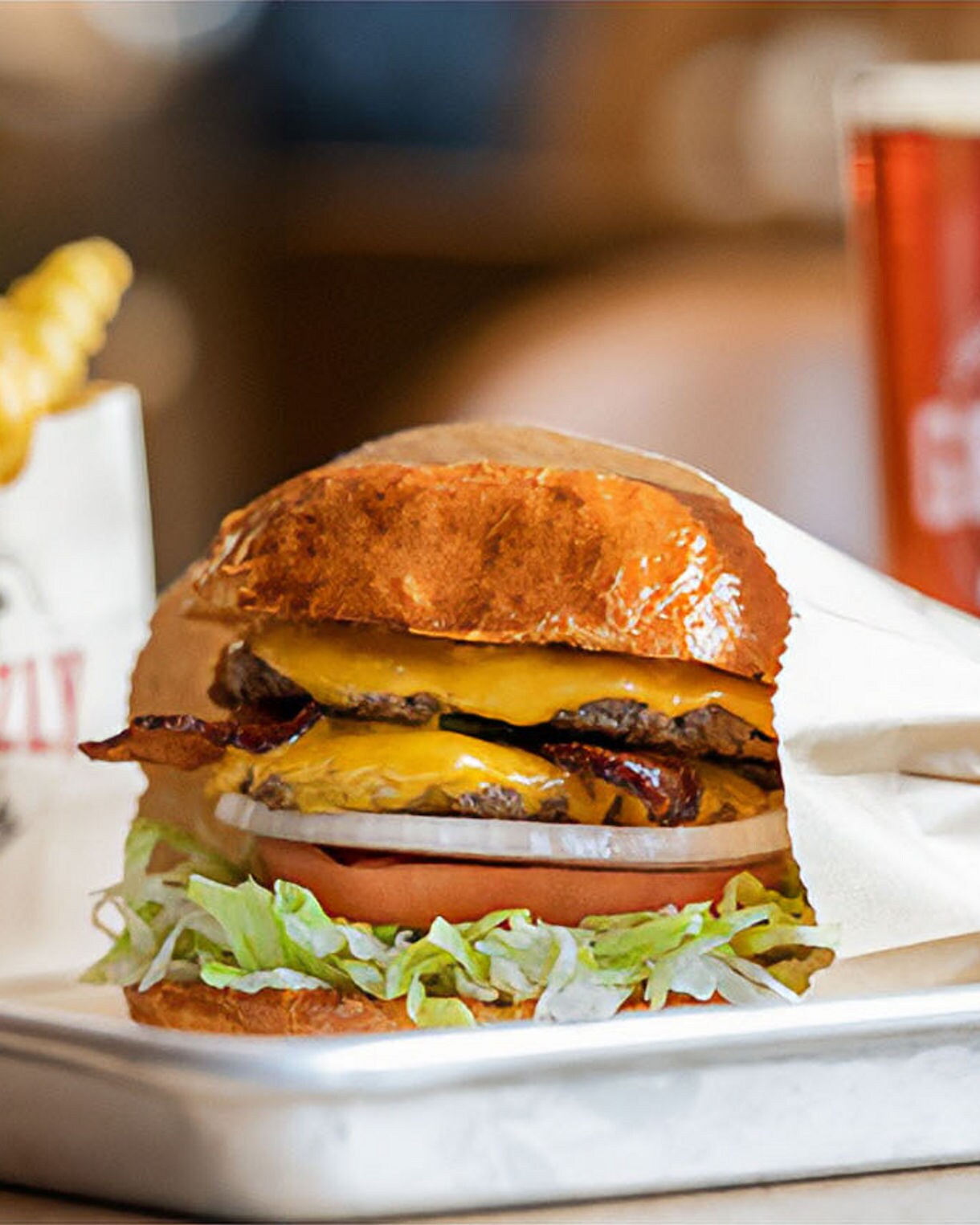 A cheeseburger with bacon, lettuce, tomato, and onion is served on a metal tray alongside crinkle-cut fries in a branded Grizzly bag. A beverage in a clear cup with the Grizzly logo is also visible in the background. The setting appears to be a casual dining restaurant with a warm, inviting atmosphere. The focus is on the fresh ingredients and appetizing presentation. The word 'GRIZZLY' is clearly visible on both the fry bag and drink cup.