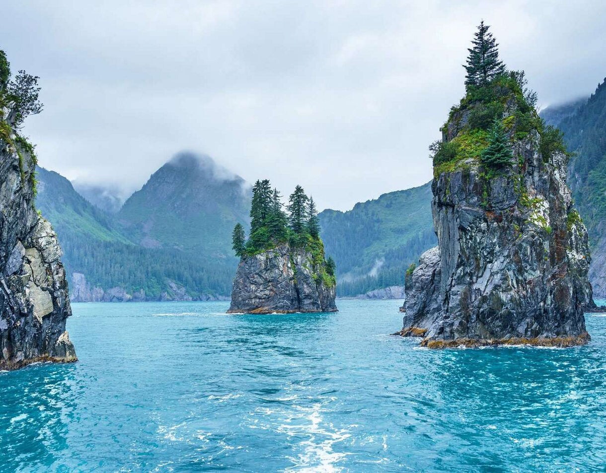Dramatic coastal rock formations with evergreen trees rising from turquoise water, shrouded in mist with forested mountains in background