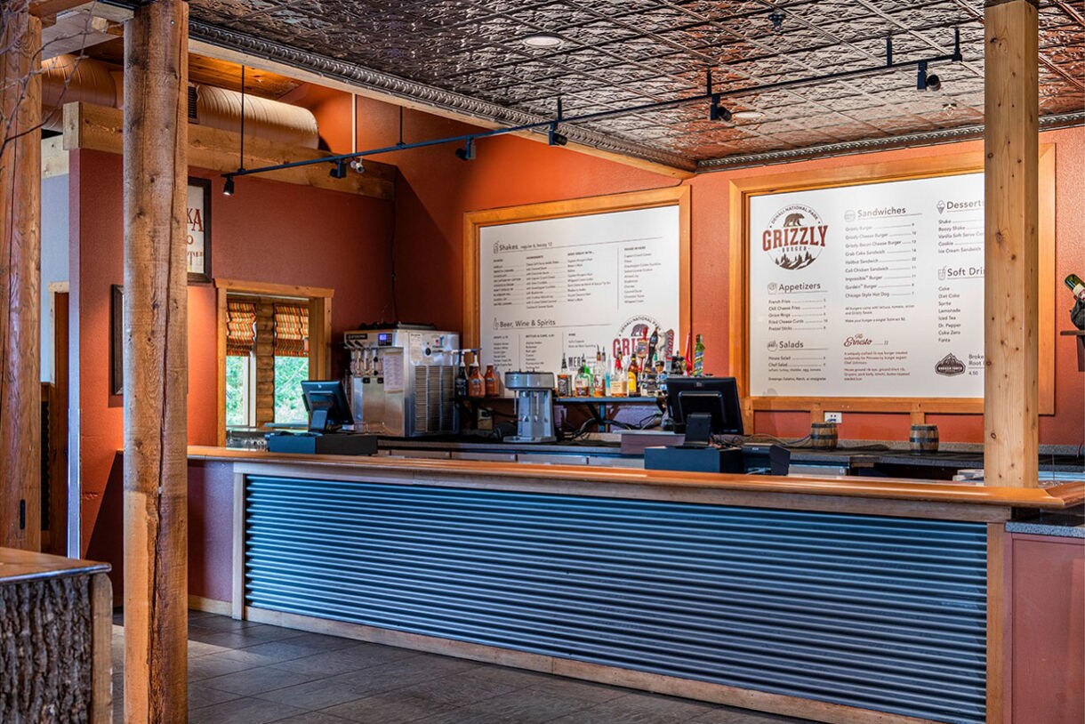 A cozy cafe counter features a corrugated metal front and wooden accents, set against a warm-toned interior. The back wall displays large menu boards with clear headings, including the word 'GRIZZLY' and various drink and food options. Bottles, a coffee machine, and a cash register are visible on the counter, suggesting a casual dining or coffee shop environment. The setting is well-lit with natural and artificial light, and the overall mood is inviting and relaxed.