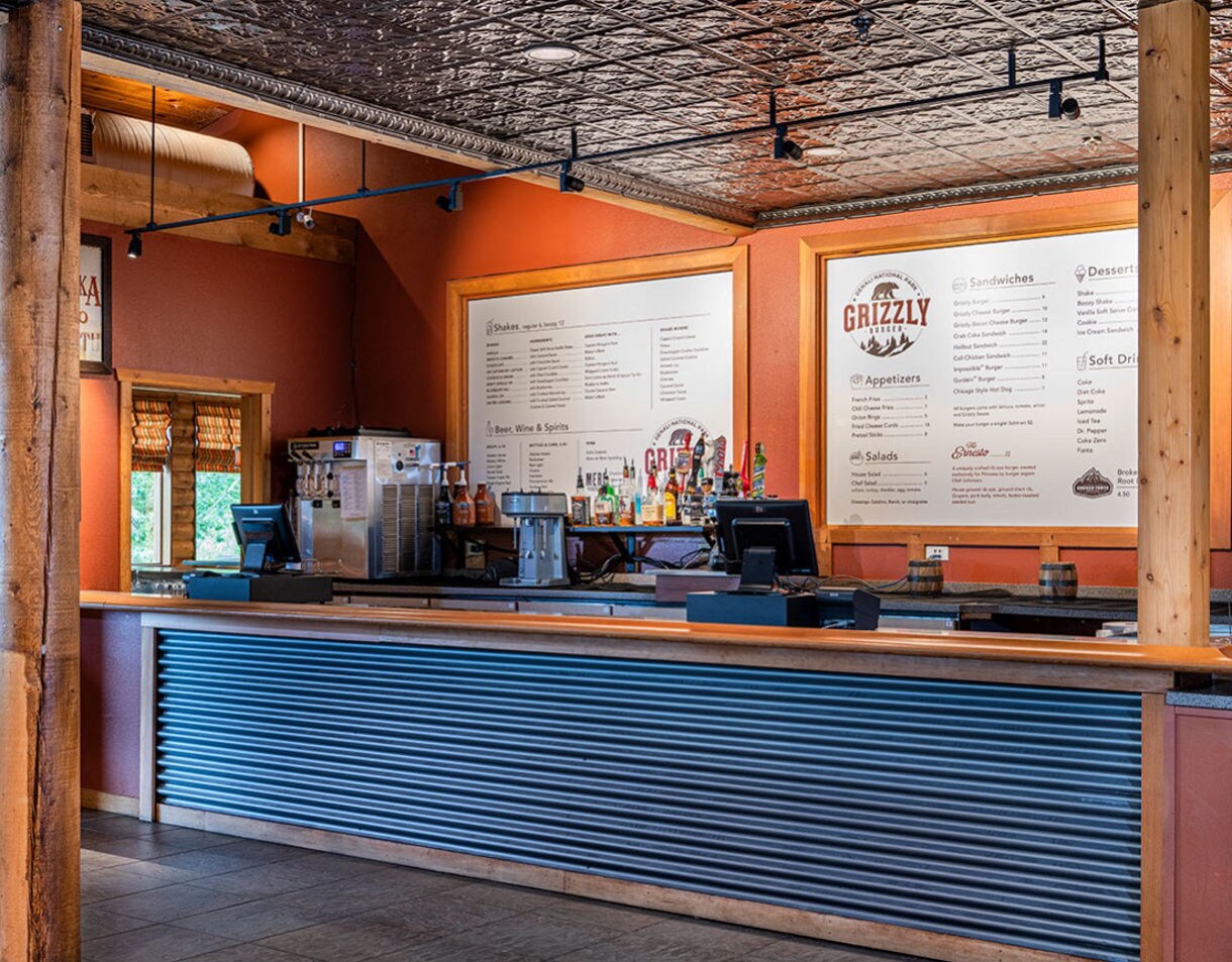 A cozy cafe counter features a corrugated metal front and wooden accents, set against a warm-toned interior. The back wall displays large menu boards with clear headings, including the word 'GRIZZLY' and various drink and food options. Bottles, a coffee machine, and a cash register are visible on the counter, suggesting a casual dining or coffee shop environment. The setting is well-lit with natural and artificial light, and the overall mood is inviting and relaxed.