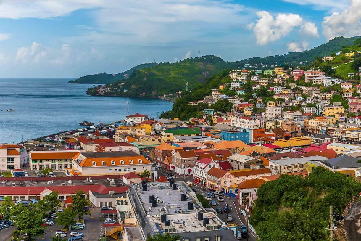 Panoramic view of St. George’s, Grenada with colorful buildings climbing a green hillside, a stone church with red rooftops in the foreground, and a calm bay stretching into the distance.