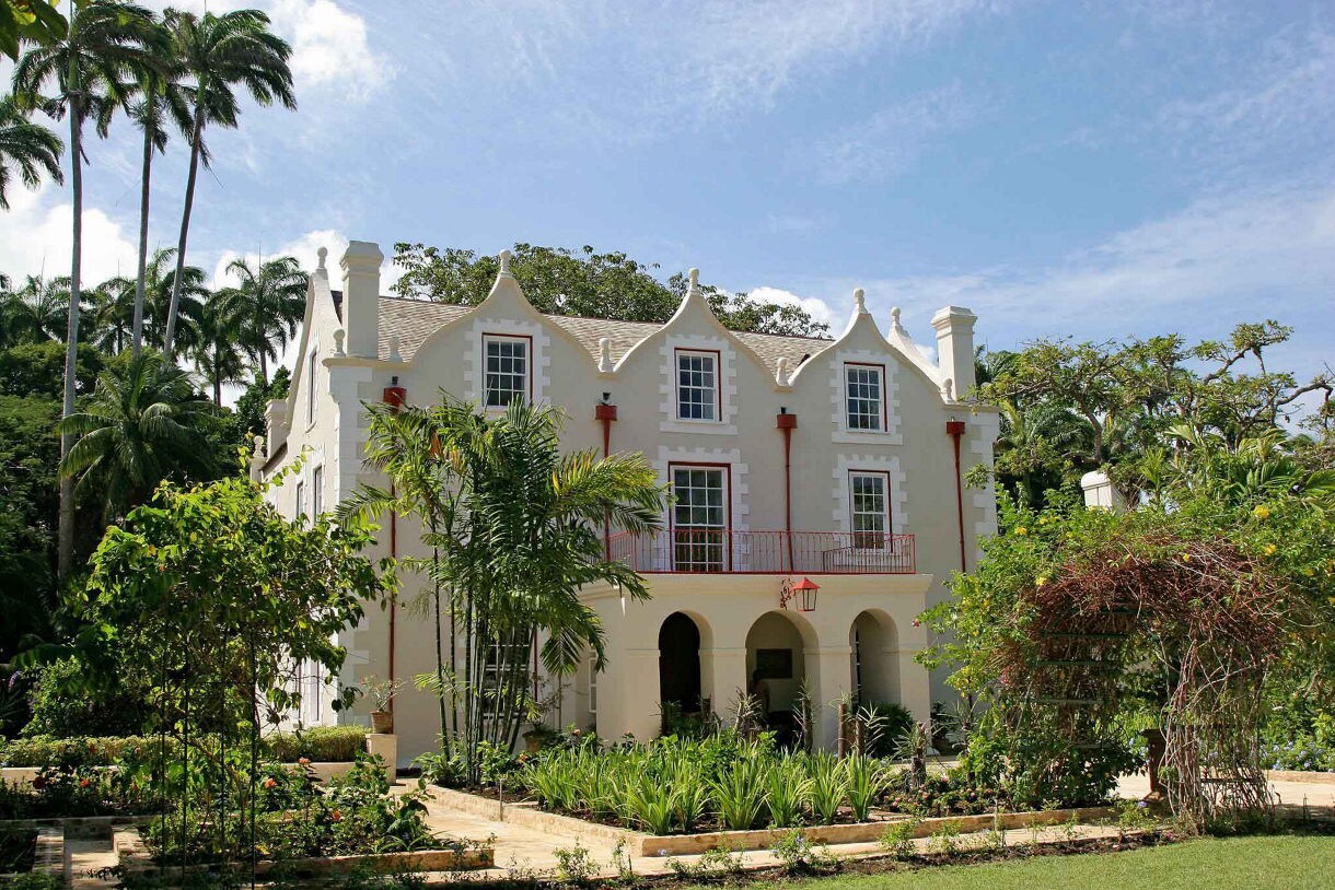 Historic St. Nicholas Abbey in Barbados featuring colonial architecture with gabled roofs, arched doorways, red trim and vibrant tropical gardens in front.
