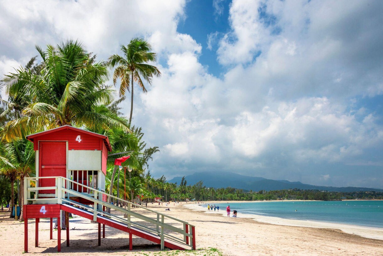 Red lifeguard tower on Luquillo Beach in Puerto Rico, with palm trees, beachgoers and distant mountains under a partly cloudy sky.