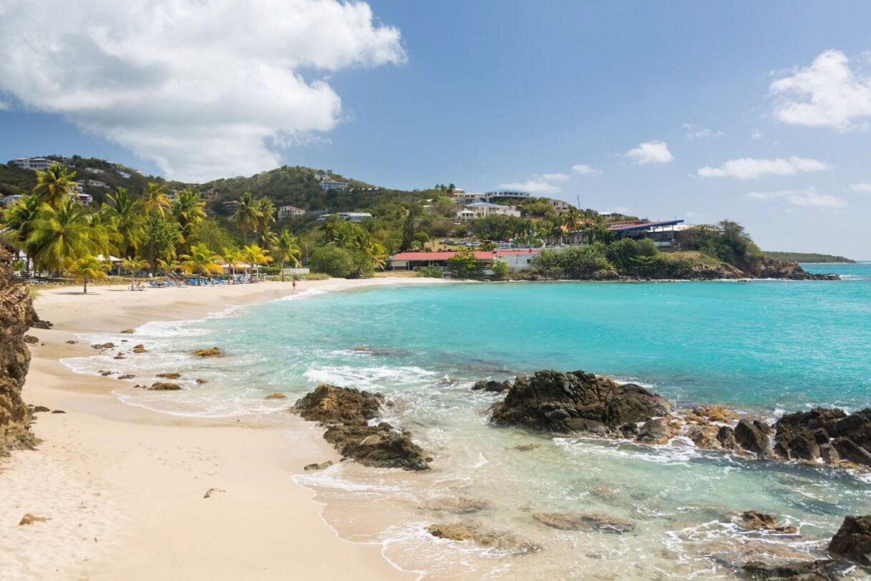 Coastal view of a beach in St. Thomas with clear turquoise water, gentle waves, palm trees, rocky outcrops and hillside buildings under a sunny sky.