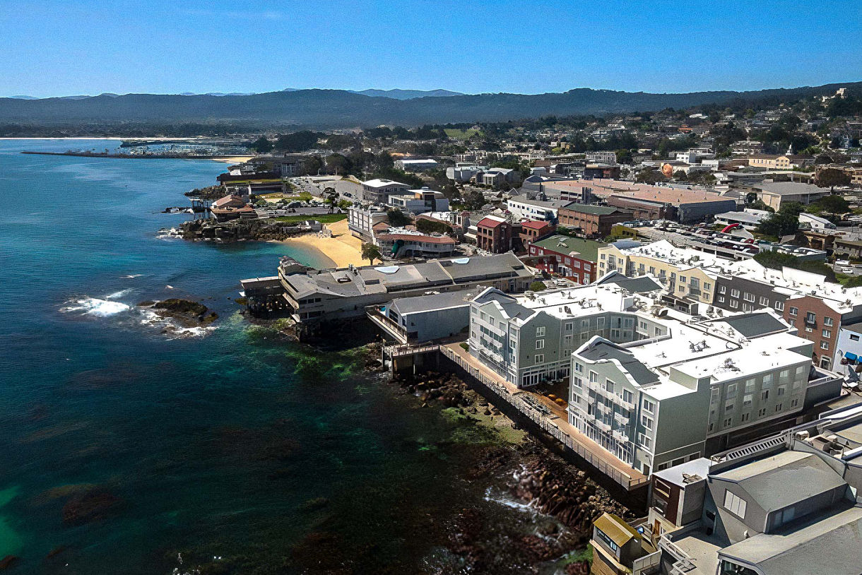 Aerial view of a coastal town with buildings along the shoreline, a small beach, and mountains in the background under a clear blue sky.