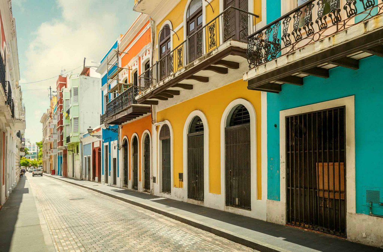 A quiet street in Old San Juan featuring colorful colonial buildings with arched windows, wrought-iron balconies and a cobblestone road under a sunny sky.