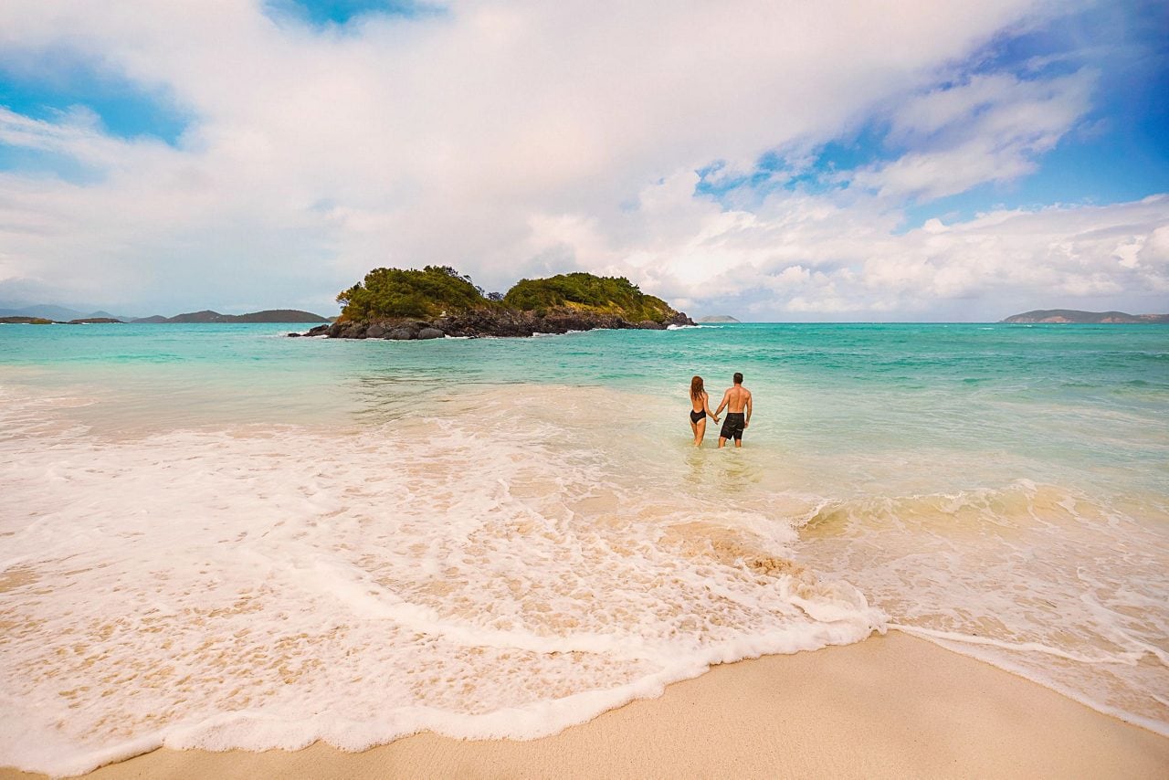 A couple walks into turquoise ocean waters on a tropical white-sand beach.