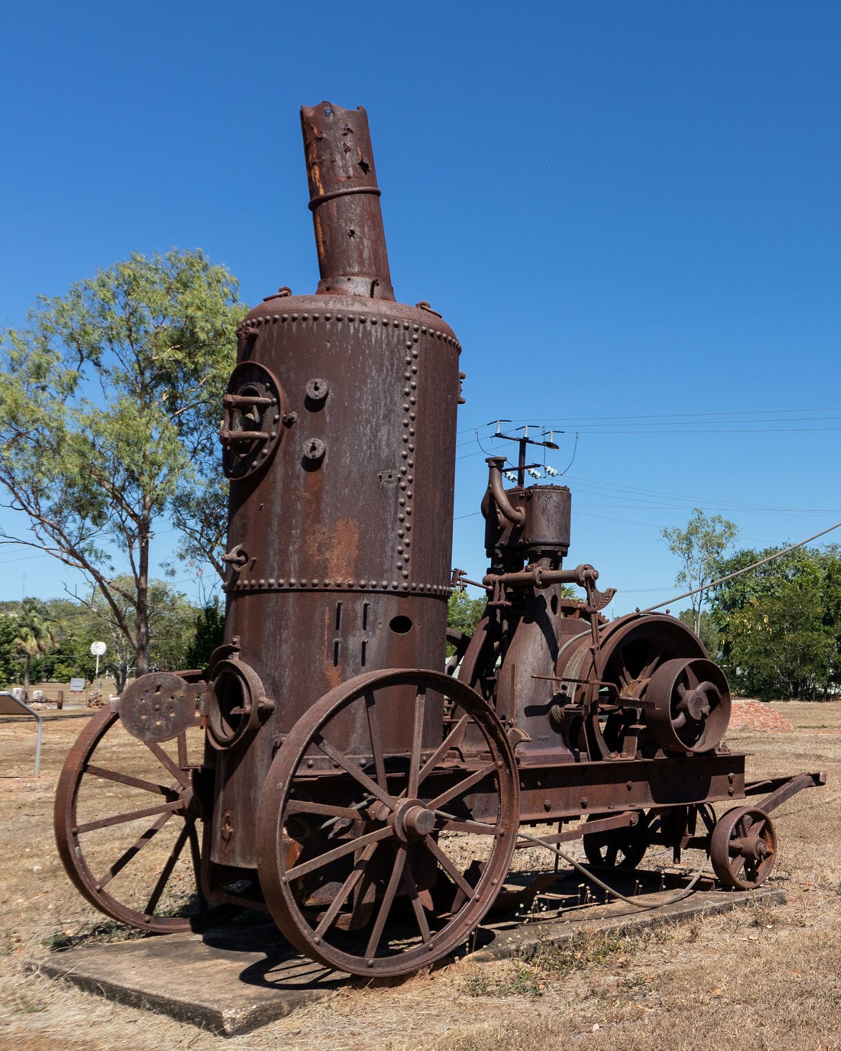 Rusted steam machinery and a tall metal headframe in a dry open field under a clear blue sky.