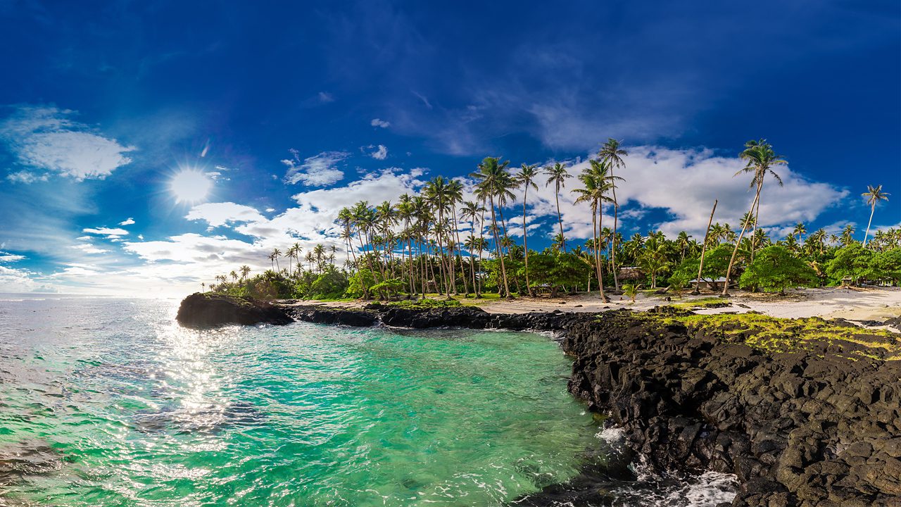 Panoramic view of a tropical beach with clear turquoise waters, palm trees, and black volcanic rocks under a sunny blue sky.