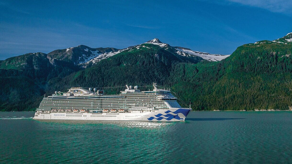  A Princess Cruises ship anchored in a calm inlet with heavily forested mountains and glacial peaks reflected in the water.