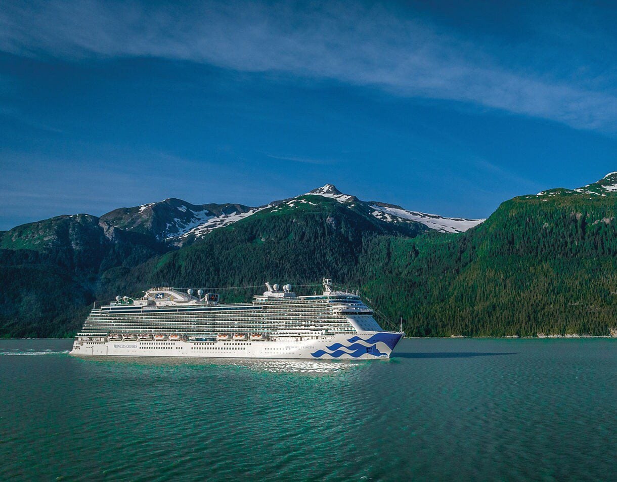 A Princess Cruises ship anchored in a calm inlet with heavily forested mountains and glacial peaks reflected in the water.