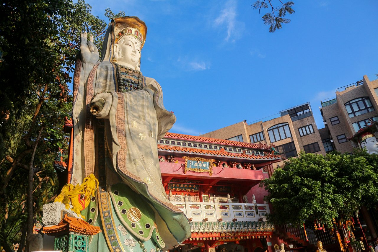 Large statue of the sea goddess outside A-Ma Temple, surrounded by colorful architectural details and red-roofed buildings under a bright blue sky.