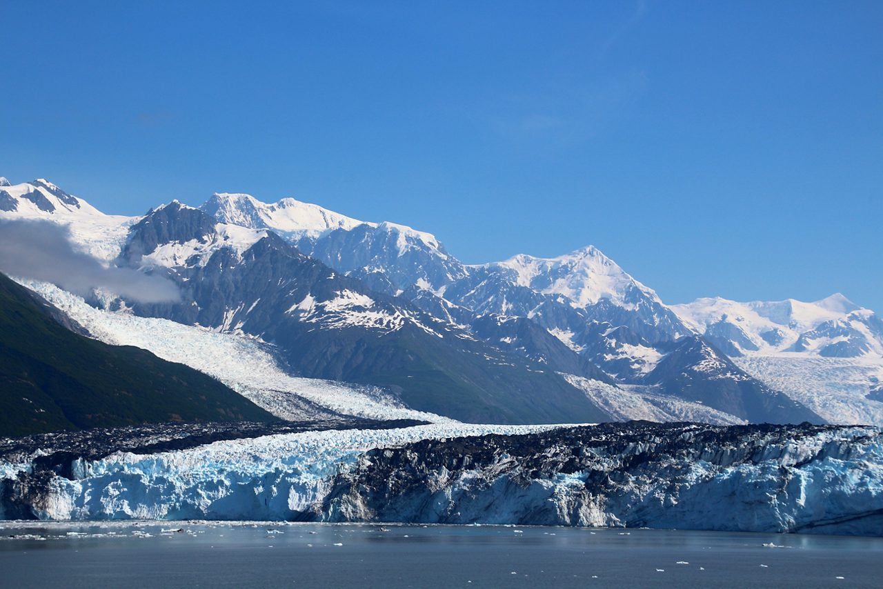 A sweeping view of College Fjord in Alaska, featuring massive glaciers flowing down from snow-capped mountains into the calm waters below.