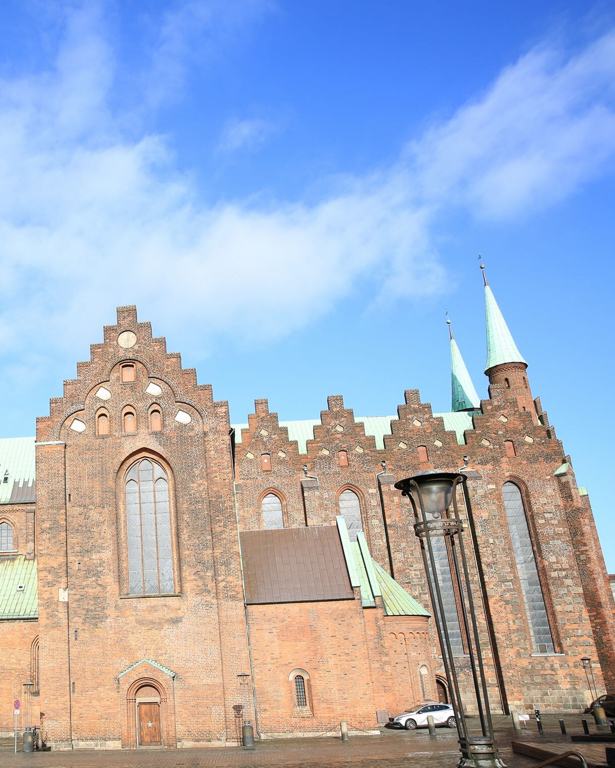 Exterior of Aarhus Cathedral with red brick walls, tall arched windows and a green copper spire under blue sky.