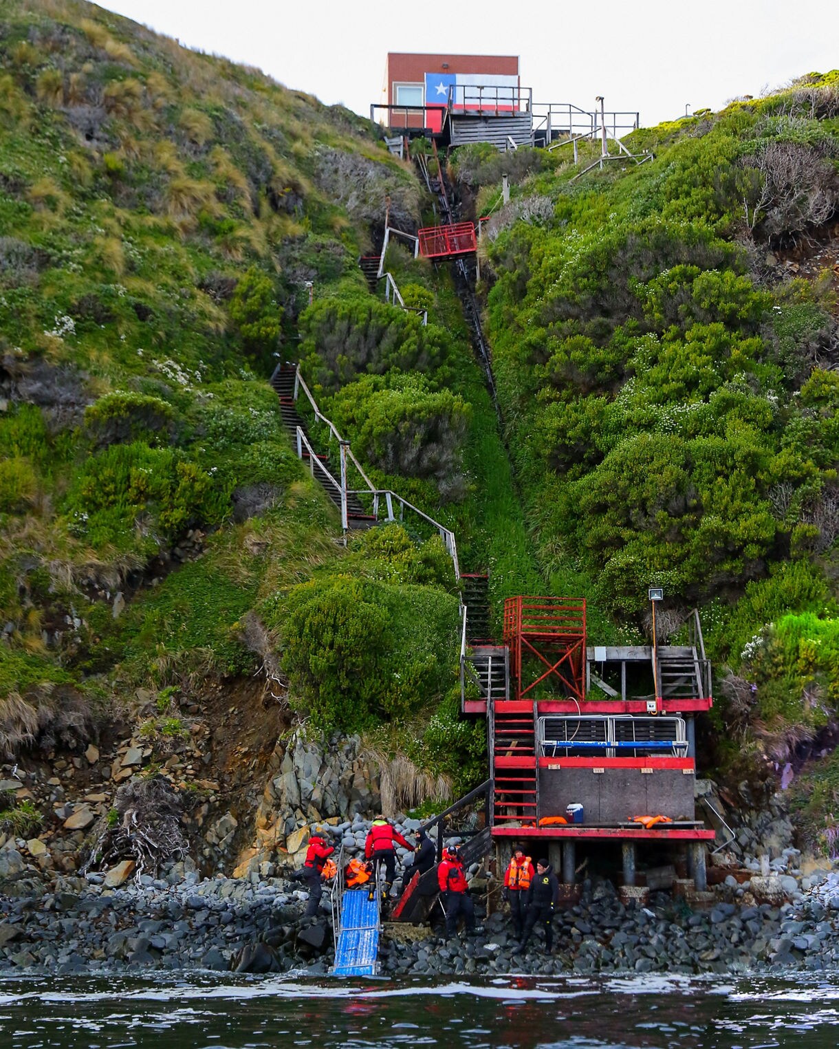 Travelers in red jackets ascending a rugged metal staircase from rocky shoreline to a steep green hillside that leads up to a small Chilean outpost.