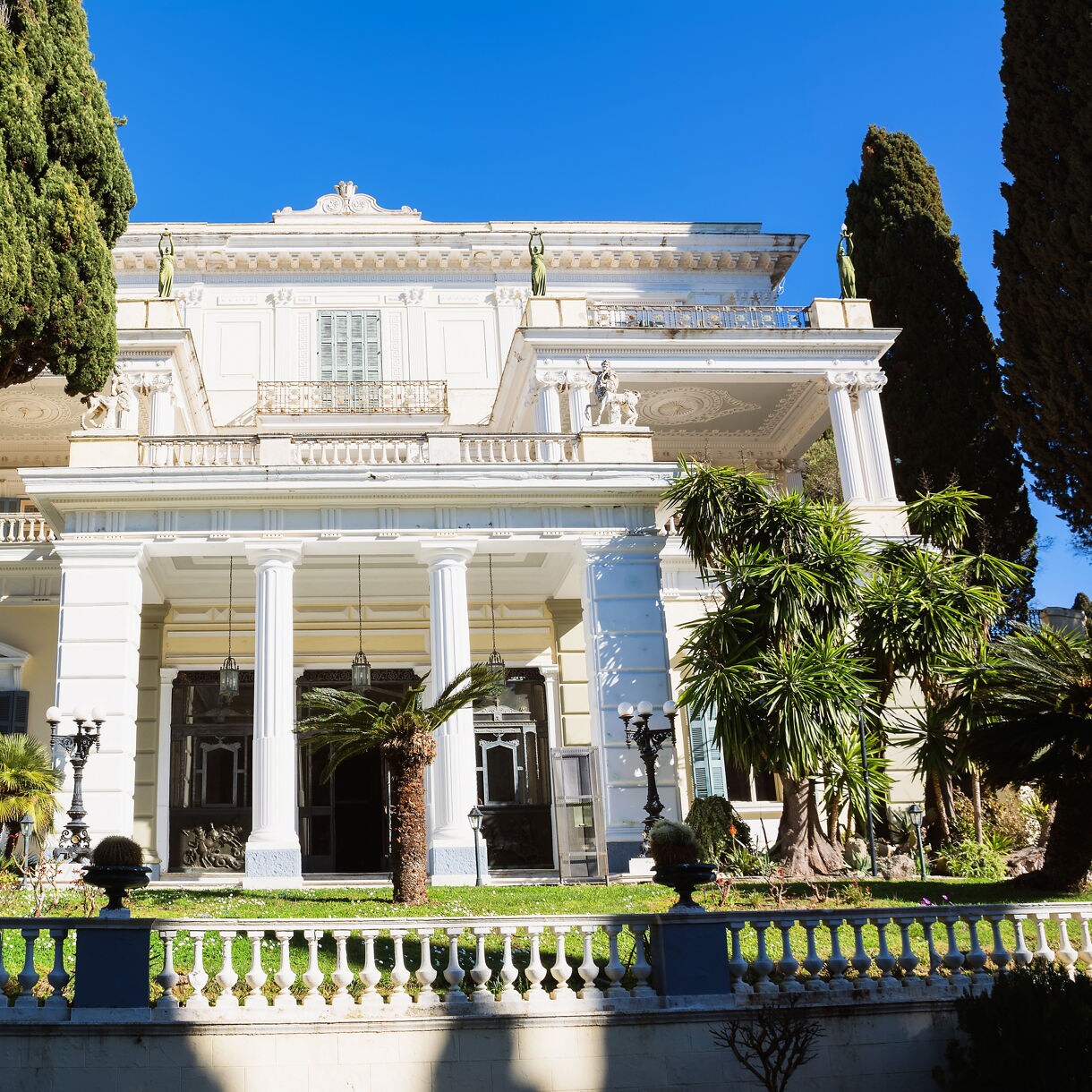 Achilleion Palace in Corfu, a grand neoclassical mansion with tall white columns, lush gardens and ornate statues.