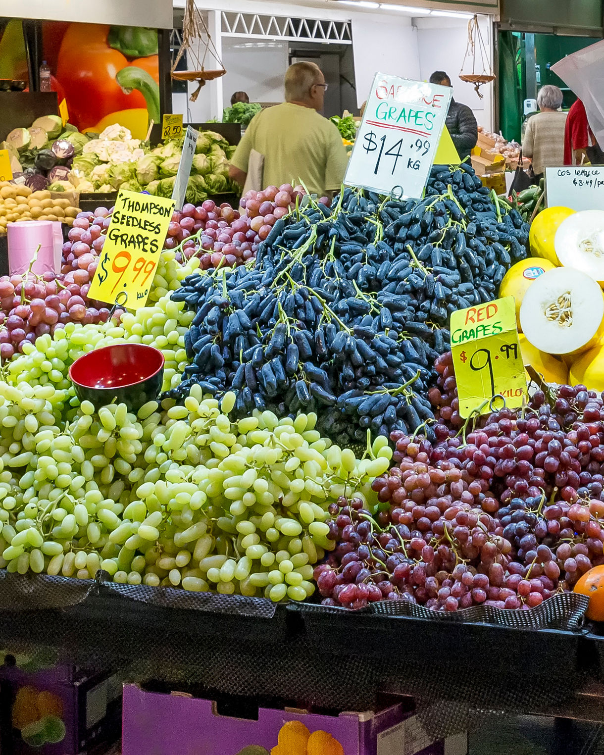 Vibrant display of grapes, melons, persimmons and avocados at Adelaide Central Market with price signs and shoppers in the background.
