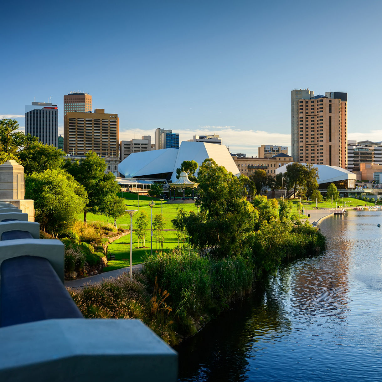 Adelaide cityscape at sunset featuring tall buildings, green riverside parks and the River Torrens with a fountain spraying water.