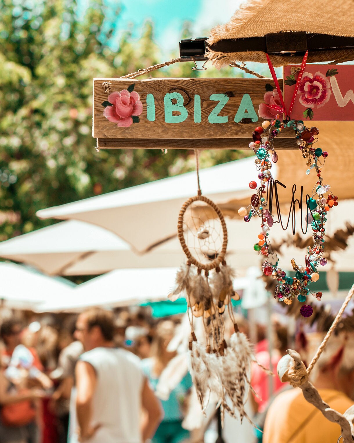 Colorful outdoor market in Ibiza with people browsing stalls and handmade crafts hanging under sunny umbrellas.