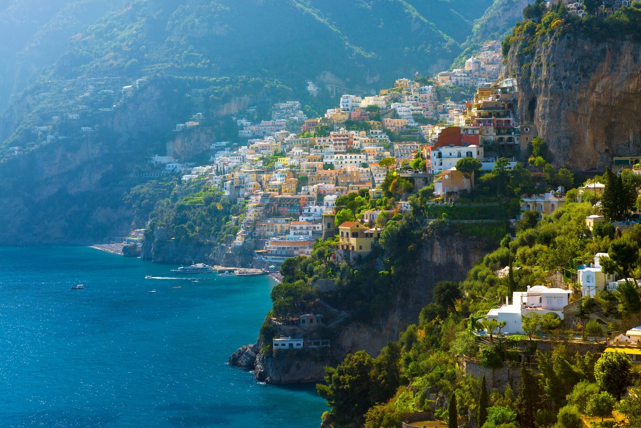 Colorful cliffside town on the Mediterranean coast with terracotta rooftops, a church tower, and lemons hanging in the foreground, backed by rugged mountains and bright blue sea.