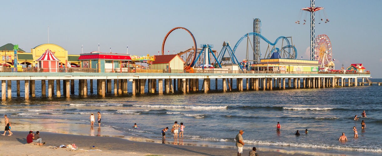 Aerial view of Galveston's Pleasure Pier, a colorful amusement pier with a roller coaster and Ferris wheel extending over the Gulf of Mexico, with beachgoers on the sand below.