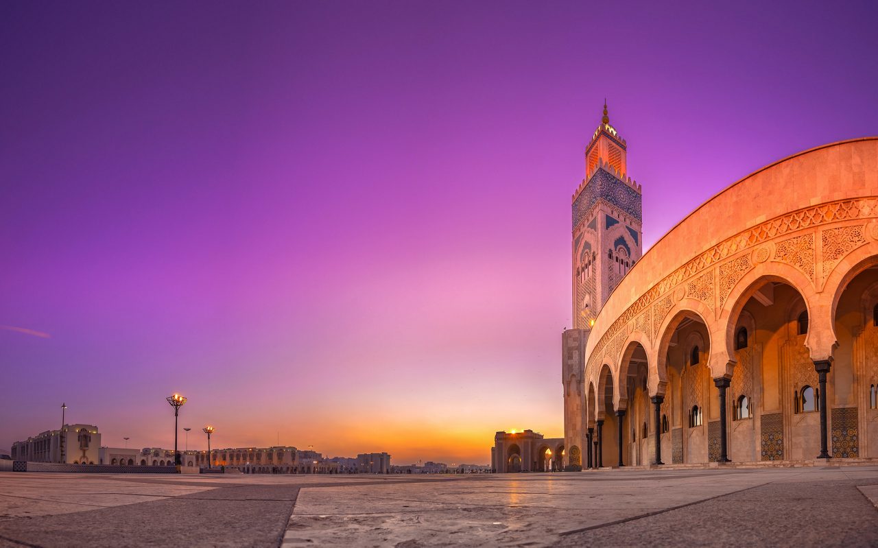Stone archways with green decorative tiles open onto a wide plaza with city buildings in the distance under a bright sky.