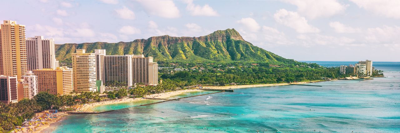 Waikiki Beach and high-rise hotels curving along the shoreline beneath the green slopes of Diamond Head, with turquoise water in the foreground.