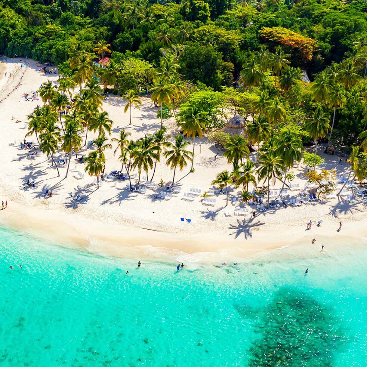 Aerial drone view of a beautiful Caribbean tropical island featuring white sandy beaches, turquoise water, lush palm trees and people enjoying the serene shoreline.