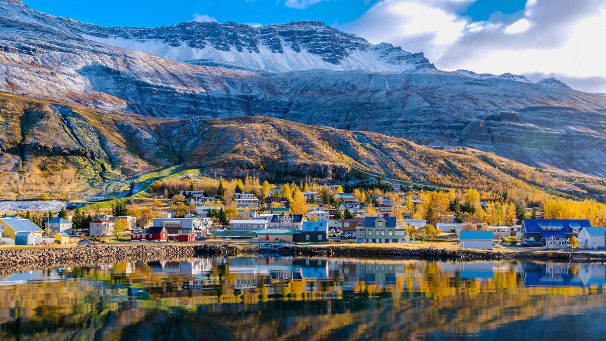 A small coastal town with colorful houses along a calm bay, reflected in glassy water and framed by snow-dusted mountains glowing in warm autumn sunlight.