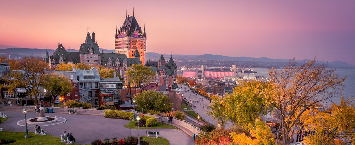 Aerial view of Quebec City at dusk, with the Château Frontenac lit up above the historic walled old town and the St. Lawrence River in the background.