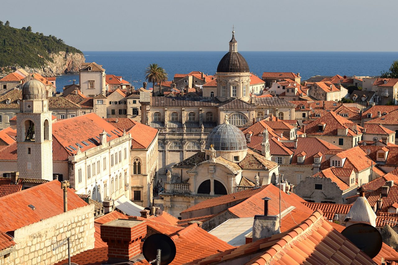 Aerial view over Dubrovnik's Old Town rooftops, showing terracotta-tiled buildings, church domes and bell towers, with the Adriatic Sea and coastline in the background.