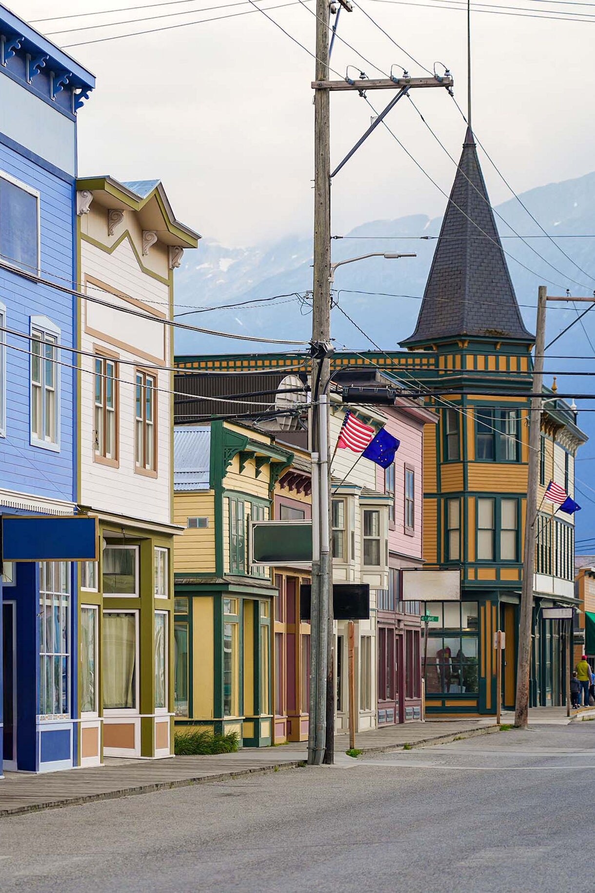 A row of colorful historic buildings lining a street in Skagway, Alaska, with flags fluttering in the breeze and a cruise ship visible in the distance.