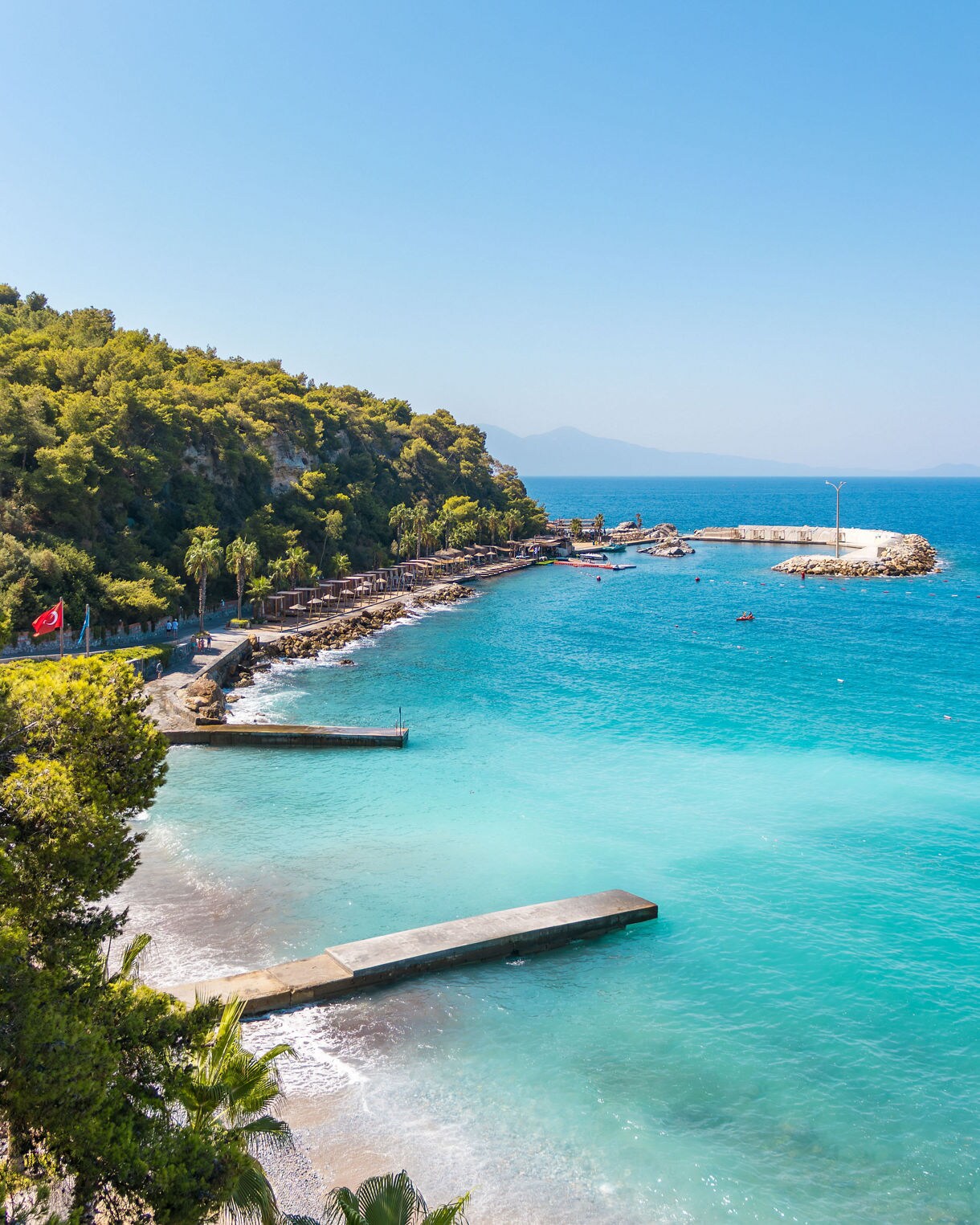 Turquoise shoreline with small piers and rocky edges, backed by green hills and open blue sea under a clear sky.
