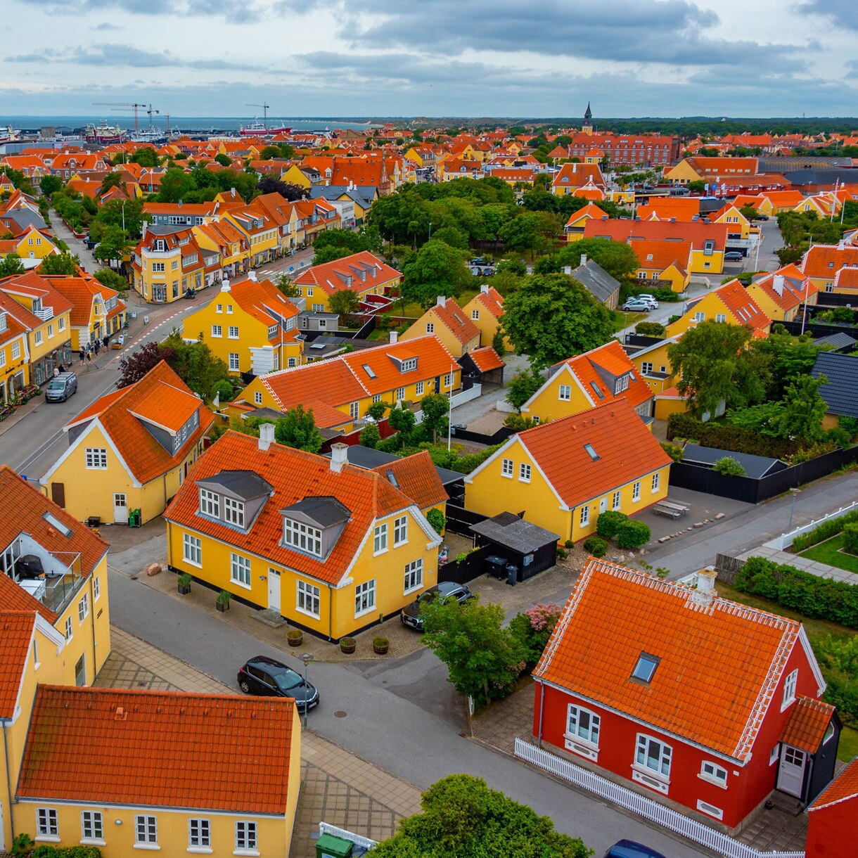 Aerial view of Skagen, Denmark, showing rows of yellow houses with red tiled roofs, green trees and the harbor in the background.